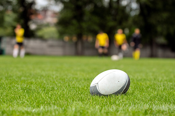 a white rugby ball on the green grass of a rugby pitch with some players wearing yellow in the distance